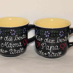 May include: Two blue ceramic mugs with yellow interiors and white lettering. The mug on the left says "F&uuml;r die beste Mama der Welt" and the mug on the right says "F&uuml;r den besten Papa der Welt".
