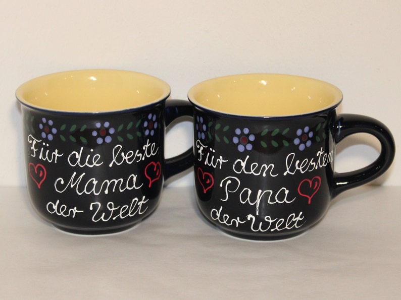 May include: Two blue ceramic mugs with yellow interiors and white lettering. The mug on the left says "F&uuml;r die beste Mama der Welt" and the mug on the right says "F&uuml;r den besten Papa der Welt".