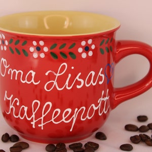 Könnte beinhalten: Rote Keramik-Kaffeetasse mit gelbem Rand und weißer Aufschrift " Oma Lisas Kaffeepott". Die Tasse hat ein grünes und weißes Blumenmuster am oberen Rand.