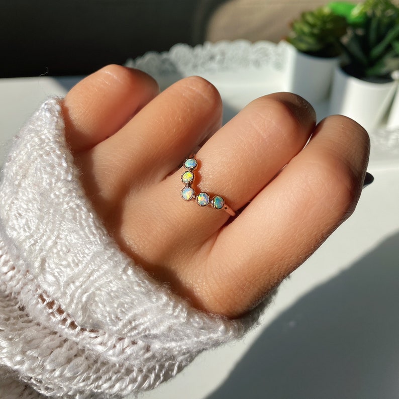 May include: A rose gold ring featuring a curved design with six opal gemstones. The opals display a range of colors, including white, yellow, and blue. The ring is worn on a finger, showcasing its delicate and elegant style.