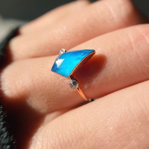 Könnte beinhalten: Ein Ring mit einem großen, facettierten, blauen Edelstein, der in einer roségoldenen Fassung sitzt. Der Edelstein ist diamantförmig und hat eine spitze Spitze. Auf der Fassung befinden sich zwei kleinere, klare Edelsteine.