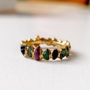 May include: A gold band ring with a row of colorful gemstones. The ring features green, black, and pink stones set in a gold band.