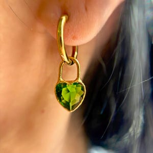 May include: Gold hoop earrings with a green heart-shaped charm. The charm is shaped like a padlock with a heart-shaped keyhole.