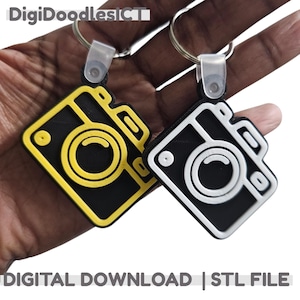 May include: Two camera-shaped keychains. One is black with a yellow outline, and the other is black with a white outline. Both have a circular lens design and are attached to a silver keyring. The text "DIGITAL DOWNLOAD | STL FILE" is at the bottom.