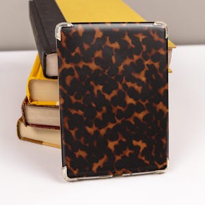 May include: A rectangular case with a tortoiseshell pattern, featuring dark brown and amber tones, rests against a stack of books. The case has clear corner protectors. The books have yellow, black, and red covers.