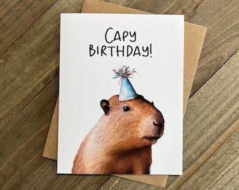 May include: A white card with the text "Capy Birthday!" in black marker. A capybara wearing a blue and white party hat is pictured on the card.