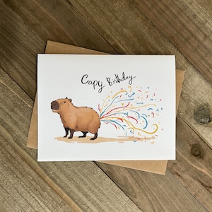 May include: A white card with a cartoon capybara with a stream of colorful confetti coming out of its rear end. The card says "Capy Birthday" in black cursive writing.