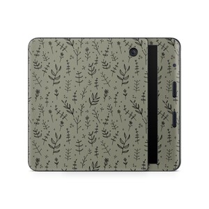 May include: An e-reader device featuring a sage green skin with a repeating black line art pattern of small flowers, leaves, and stems. The device has a black screen bezel, a circular camera lens on the back, and visible side control buttons.