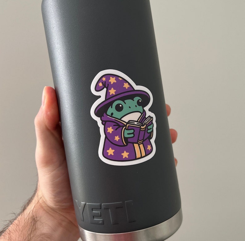 May include: A black Yeti water bottle with a sticker of a green frog wearing a purple wizard hat and holding a book. The frog has a yellow star on its hat and a yellow star on its robe.