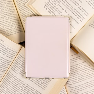 May include: A light pink tablet or e-reader device with a clear protective case, resting on open books. The device is rectangular with rounded corners, suggesting a modern design, and is surrounded by pages of text.