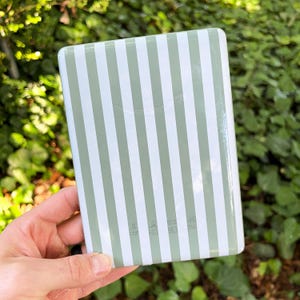 May include: A rectangular e-reader device featuring a vertical striped pattern in sage green and white. The back of the device displays a subtle embossed logo near the top center and small regulatory text printed in white near the bottom edge.