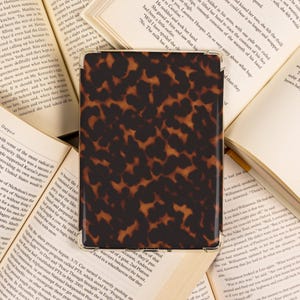 May include: A rectangular case with a tortoiseshell pattern, likely for a tablet or e-reader, rests on open books. The case features a glossy finish and a dark brown and tan color scheme, suggesting a classic design.