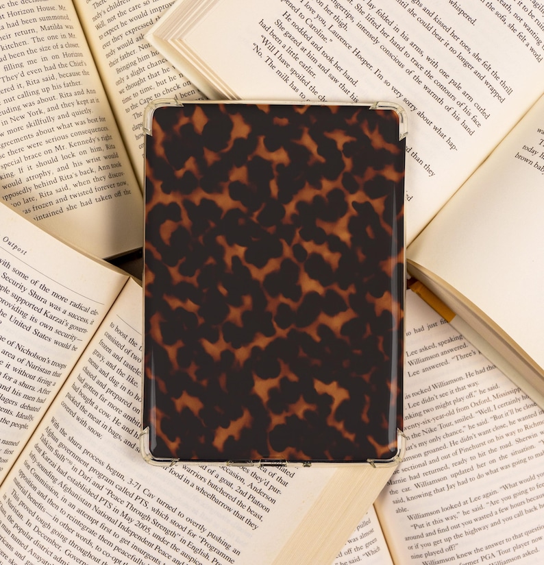May include: A rectangular case with a tortoiseshell pattern, likely for a tablet or e-reader, rests on open books. The case features a glossy finish and a dark brown and tan color scheme, suggesting a classic design.