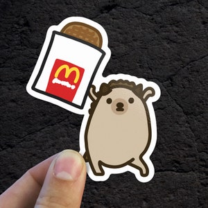 May include: A cartoon hedgehog holding a red and yellow McDonald's bag with a french fry inside. The hedgehog is brown with a white belly and has a friendly expression.