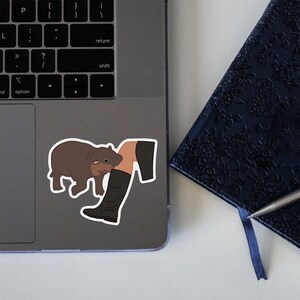 May include: A sticker featuring a cartoon illustration of a person's leg in a black boot being nibbled on by a brown and tan capybara.