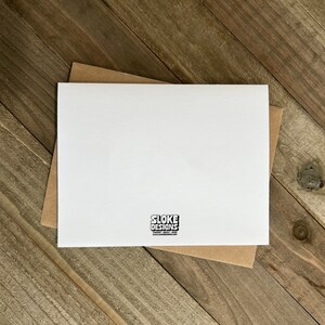 May include: Blank white card with a brown envelope. The card has a small black logo that says "Sloke Designs" with a website address.