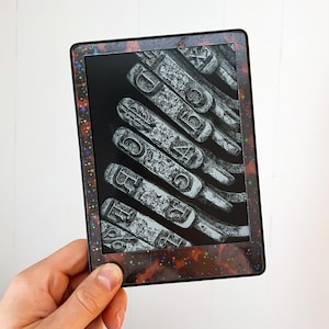 May include: A rectangular e-reader device featuring a dark, holographic frame decorated with small, colorful stars. The screen displays a high-contrast, black and white close-up image of several metallic typewriter keys arranged in a diagonal row.