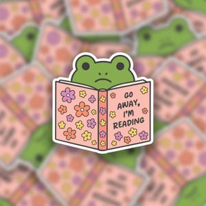 May include: A sticker featuring a green frog with a sad expression, reading a pink book. The book is decorated with colorful flowers and the text "GO AWAY, I'M READING". The background shows other stickers with the same design.