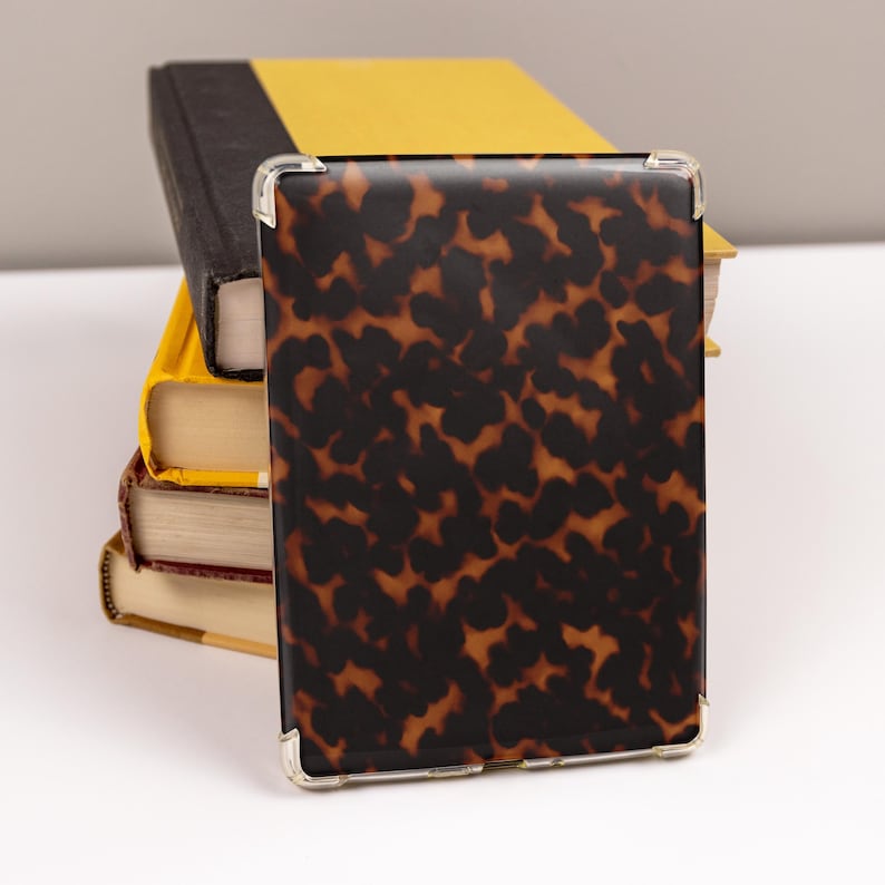 May include: A rectangular case with a tortoiseshell pattern, featuring dark brown and amber tones, rests against a stack of books. The case has clear corner protectors. The books have yellow, black, and red covers.
