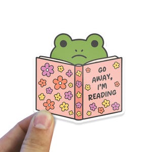 May include: A sticker featuring a green frog peeking over a pink book. The book is decorated with colorful flowers and the text "GO AWAY, I'M READING". The frog has a slightly sad expression.