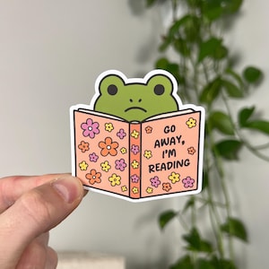 May include: A sticker featuring a green frog peeking over a book. The book is peach-colored with floral designs and the text "GO AWAY, I'M READING". The frog has a sad expression.