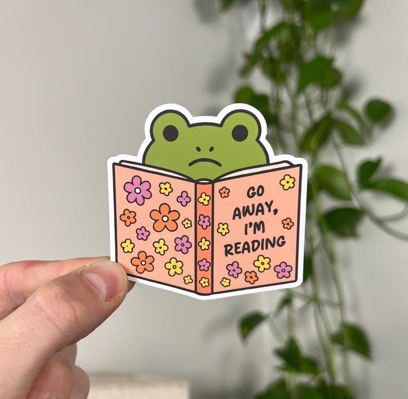 May include: A sticker featuring a green frog peeking over a book. The book is peach-colored with floral designs and the text "GO AWAY, I'M READING". The frog has a sad expression.