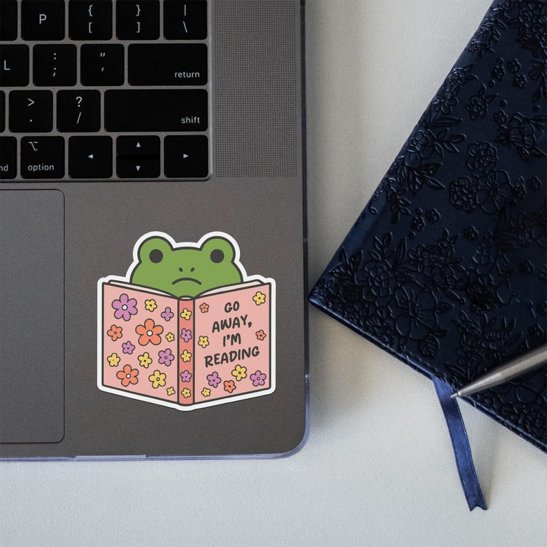 May include: A sticker featuring a green frog reading a book with the text "GO AWAY, I'M READING." The book is pink and decorated with colorful flowers. The sticker is placed on a laptop.