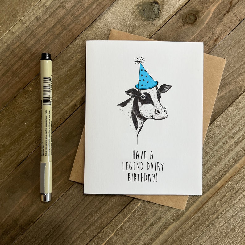 May include: A white card with a black and white illustration of a cow wearing a blue party hat. The text on the card reads "Have a Legend Dairy Birthday!"