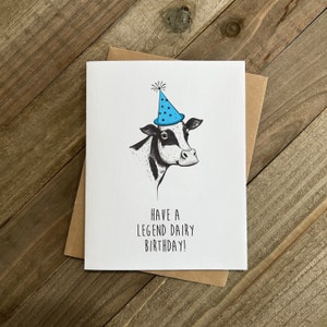 May include: A birthday card featuring a black and white cow wearing a blue party hat with white polka dots. The card reads "Have a Legend Dairy Birthday!"