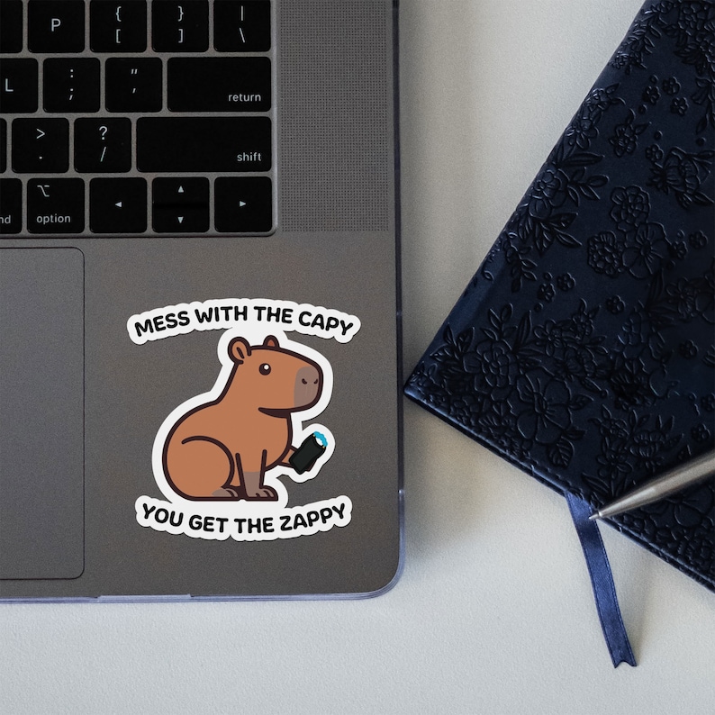 Op de afbeelding: Een sticker met een cartoon capybara die een blauwe en zwarte controller vasthoudt. De sticker zegt "Mess with the capy you get the zappy".