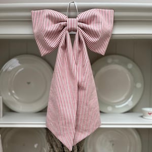 May include: A large bow made from red and white striped fabric is suspended from a white hook. The bow's fabric drapes downwards. The background shows white plates on shelves, suggesting a kitchen or dining room. The bow is a decorative item.