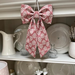 May include: A pink fabric bow with a white floral design, suspended by a white string. The bow hangs in front of a white shelf displaying white plates and a white jug. The bow is a decorative item.