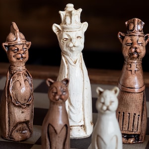 May include: A unique chess set featuring cat-shaped pieces in various colors. The pieces include a white queen with a crown, brown rooks, and pawns. The set is displayed on a checkered board, showcasing the detailed craftsmanship.