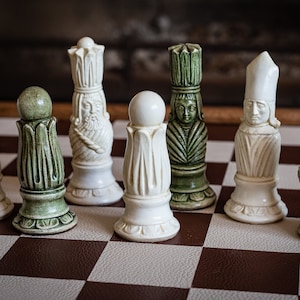 Op de afbeelding: Een set schaakstukken met groene en witte keramische ontwerpen. De stukken omvatten een toren, een paard, een loper en een pion. De stukken zijn gerangschikt op een bruin en wit geblokt schaakbord.