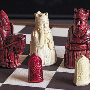 May include: Chess pieces on a checkered board. The pieces are carved in red and cream colors. The red pieces include a knight on horseback, a king, and a rook. The cream pieces include a queen and a rook.