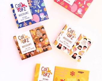 Puede incluir: Cuatro cajas de materiales artísticos de cera de abeja. Las cajas son amarillas, marrones, azules y rojas con ilustraciones coloridas de abejas, flores y personas. Las cajas están etiquetadas como "Beecolorz" y contienen crayones de cera de abeja, láminas de modelado y crayones de cera de abeja.