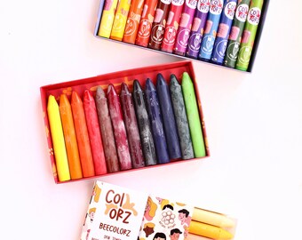 Puede incluir: Dos cajas de lápices de cera de abejas. La caja superior tiene 12 lápices en tonos de rojo, naranja, amarillo, verde, azul y morado. La caja inferior tiene 6 lápices en tonos de marrón, naranja y amarillo. Las cajas tienen ilustraciones coloridas y el texto "Col Orz Beecolorz Skin Tones Beeswax Stick Crayons".