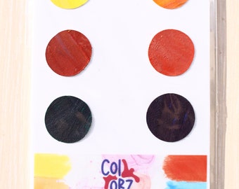 Puede incluir: Un conjunto de seis muestras de pintura de acuarela hechas a mano en amarillo, naranja, rojo, marrón, verde y morado. Las muestras de pintura están dispuestas en dos filas de tres. Las muestras de pintura están etiquetadas "Colorz Handmade Watercolorz Serie I Primary Vibrant Colorz".