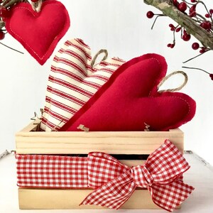 May include: Three red fabric hearts in a wooden crate with a red and white gingham ribbon bow. The hearts are decorated with white stripes and are arranged in a pile.
