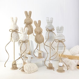 May include: Five fabric bunny figurines on wooden stands with twine bows. The bunnies are in various patterns of white, brown, and beige. The bunnies are on a white wooden surface.