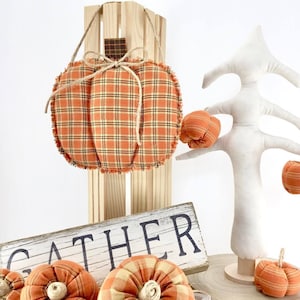May include: A wooden crate with a plaid fabric pumpkin hanging from it. The crate has the word "gather" written on it. There is a white fabric tree with three small pumpkins on it.