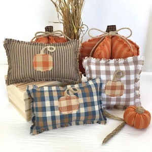 May include: A collection of fall-themed decorative pillows and pumpkins. The pillows feature various patterns, including stripes, gingham, and plaid, with a pumpkin applique. The pumpkins are orange and brown, with twine accents. The overall color scheme is warm and autumnal.