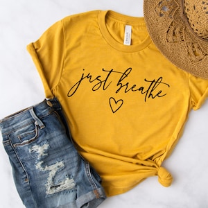 May include: A mustard yellow t-shirt with the text "just breathe" in black cursive lettering and a small heart symbol. The shirt is knotted at the bottom and is paired with denim shorts.