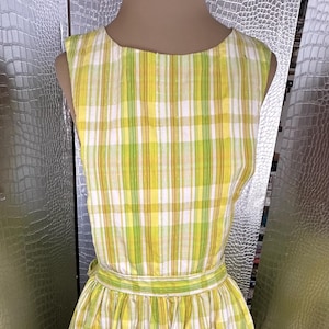 May include: Sleeveless yellow, green, and white plaid dress. The dress features a fitted bodice and a flared skirt, with a matching belt at the waist. The fabric has a vertical plaid pattern. The dress is displayed on a mannequin.