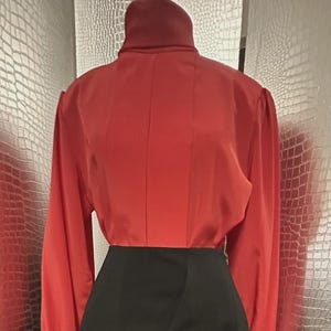 May include: A two-piece outfit featuring a long-sleeved, red blouse with a high collar and a black, wrap-style skirt. The blouse has a fitted silhouette, and the skirt is knee-length. The outfit is displayed on a mannequin.