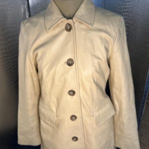 May include: A cream-colored leather jacket with a button-up front and long sleeves. The jacket has a classic design and is in good condition.