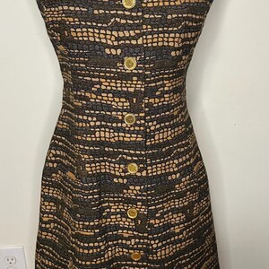 May include: A sleeveless dress featuring a textured black and tan woven pattern with a notched collar and a front button closure. The garment is secured with six round gold-toned buttons running vertically down the center of the bodice.