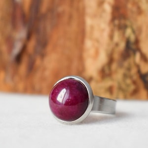 Op de afbeelding: Een roestvrijstalen ring met een grote, ronde, gepolijste, dieprode edelsteen. De edelsteen is gezet in een zilveren bezel en de ringband is een eenvoudig, geborsteld metalen ontwerp. De achtergrond is een vage houten ondergrond.