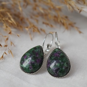 Op de afbeelding: Oorhangers in druppelvorm met een groene en paarse steen, gezet in zilverkleurig metaal. De oorbellen hebben een oorhaaksluiting. De achtergrond is een zachte witte stof.