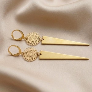 Op de afbeelding: Een paar goudkleurige oorbellen met een cirkelvormig filigraan medaillon verbonden aan een lange, gladde, driehoekige hanger. Elke oorbel is bevestigd aan een ronde klapsluiting, wat zorgt voor een gebalanceerd en geometrisch ontwerp.
